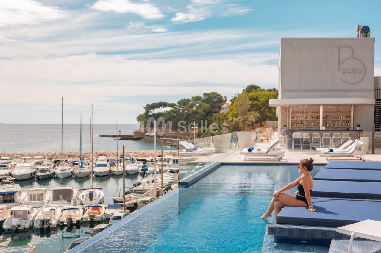 Une femme assise au bord d'une piscine surplombant un port de plaisance avec des bateaux et une vue sur la mer.