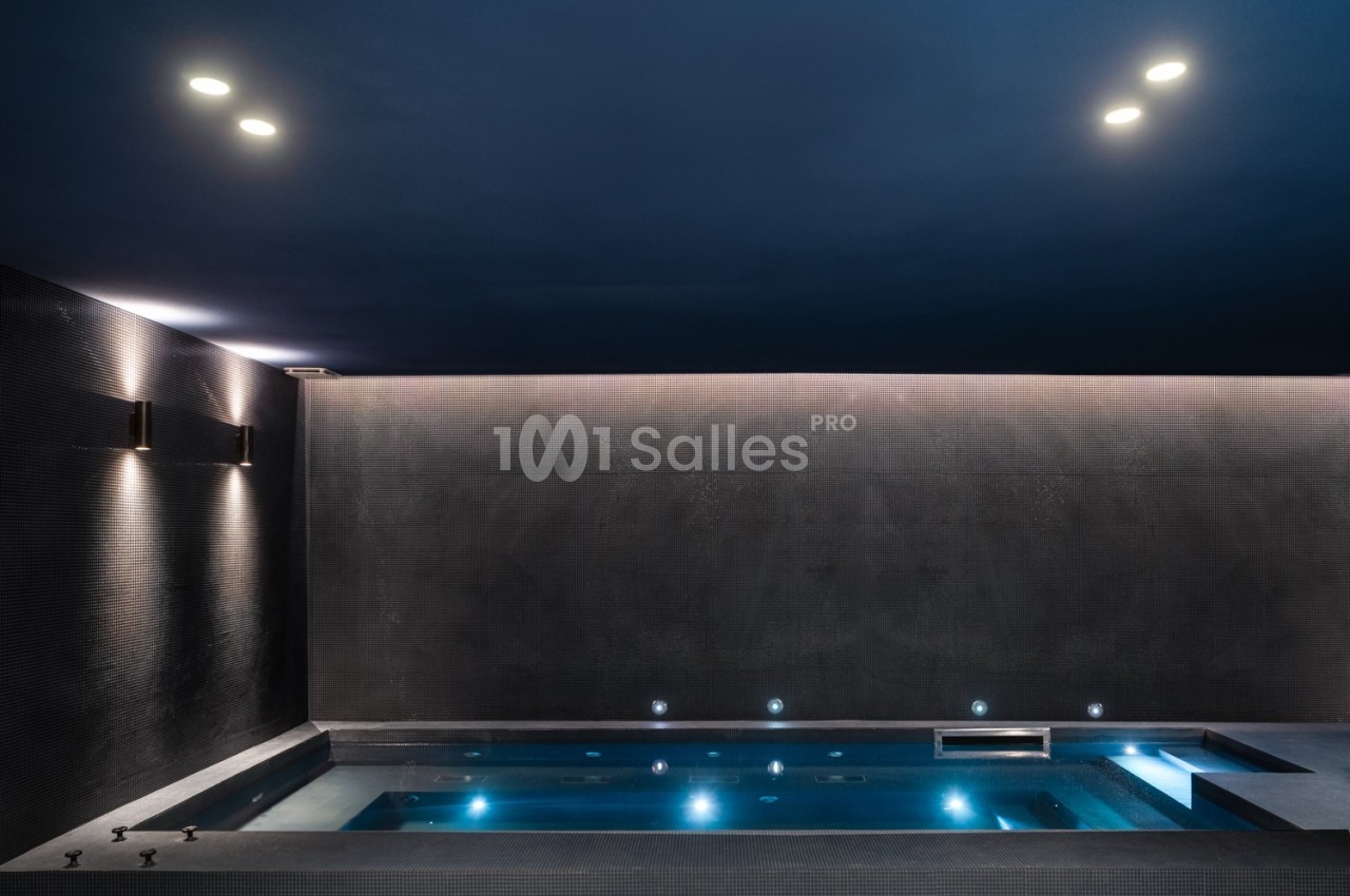 Piscine intérieure moderne éclairée par des lumières tamisées, avec un plafond sombre et des murs texturés.