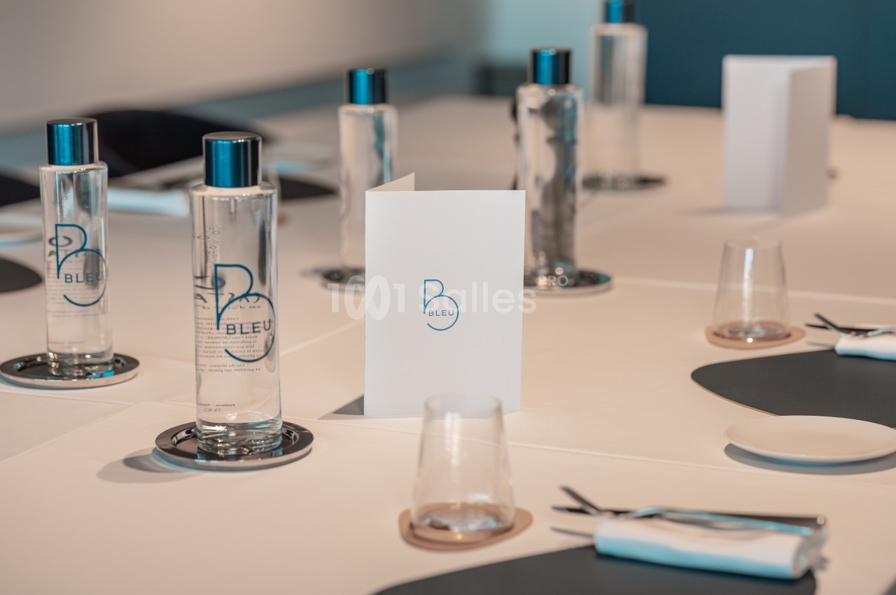Table dressée avec des bouteilles d'eau, des verres, des couverts et un menu, dans une salle lumineuse et moderne.