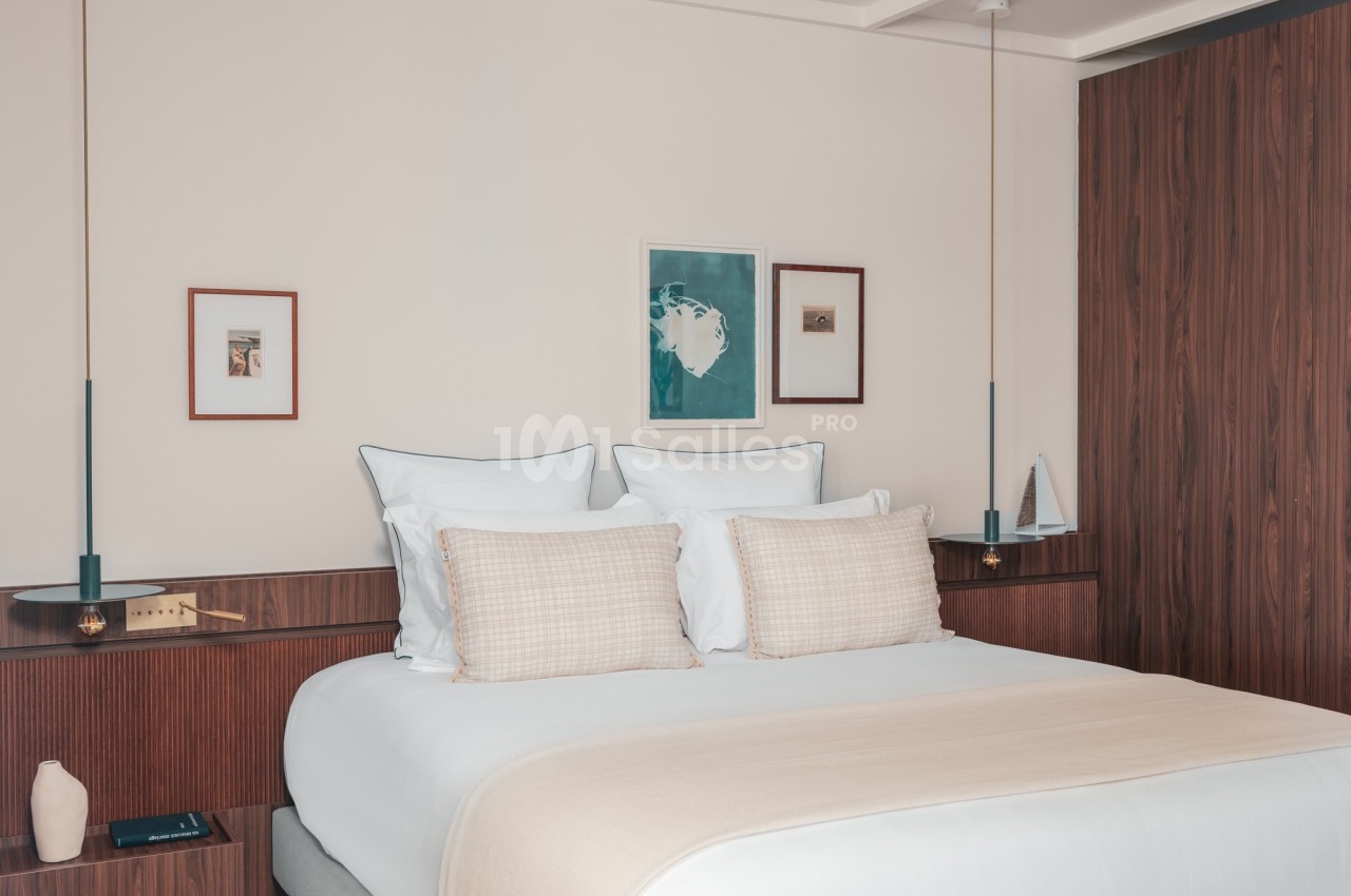 Lit double avec draps clairs, coussins beiges, tables de chevet en bois, décor minimaliste et cadres accrochés au mur.
