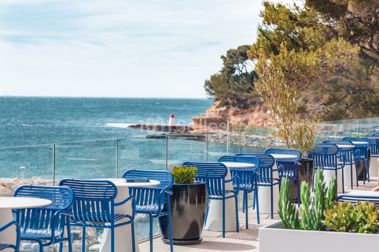 Terrasse avec tables et chaises bleues, vue sur la mer, un phare rouge et des arbres en arrière-plan.