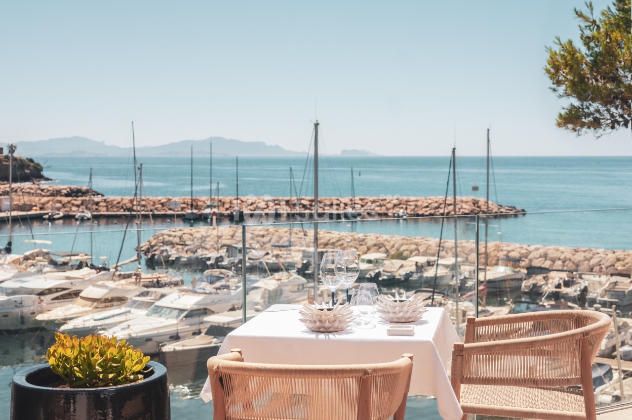 Table dressée pour deux avec vue sur un port de plaisance, des voiliers et la mer sous un ciel dégagé.