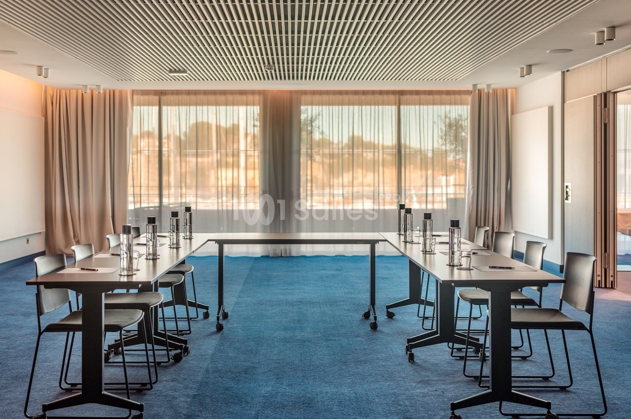 Salle de réunion lumineuse avec tables en U, chaises modernes et grandes baies vitrées donnant sur l'extérieur.