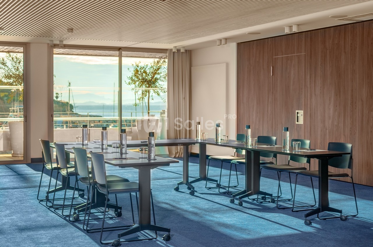 Salle de réunion lumineuse avec tables disposées en U, chaises modernes et vue sur un paysage extérieur ensoleillé.