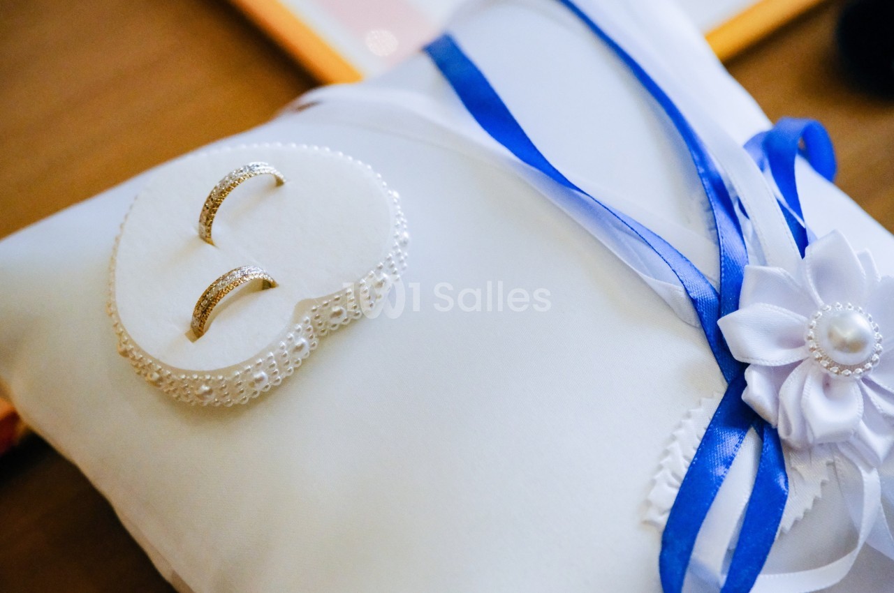 Deux alliances dorées posées sur un coussin blanc décoré de rubans bleus et d'une fleur en tissu.