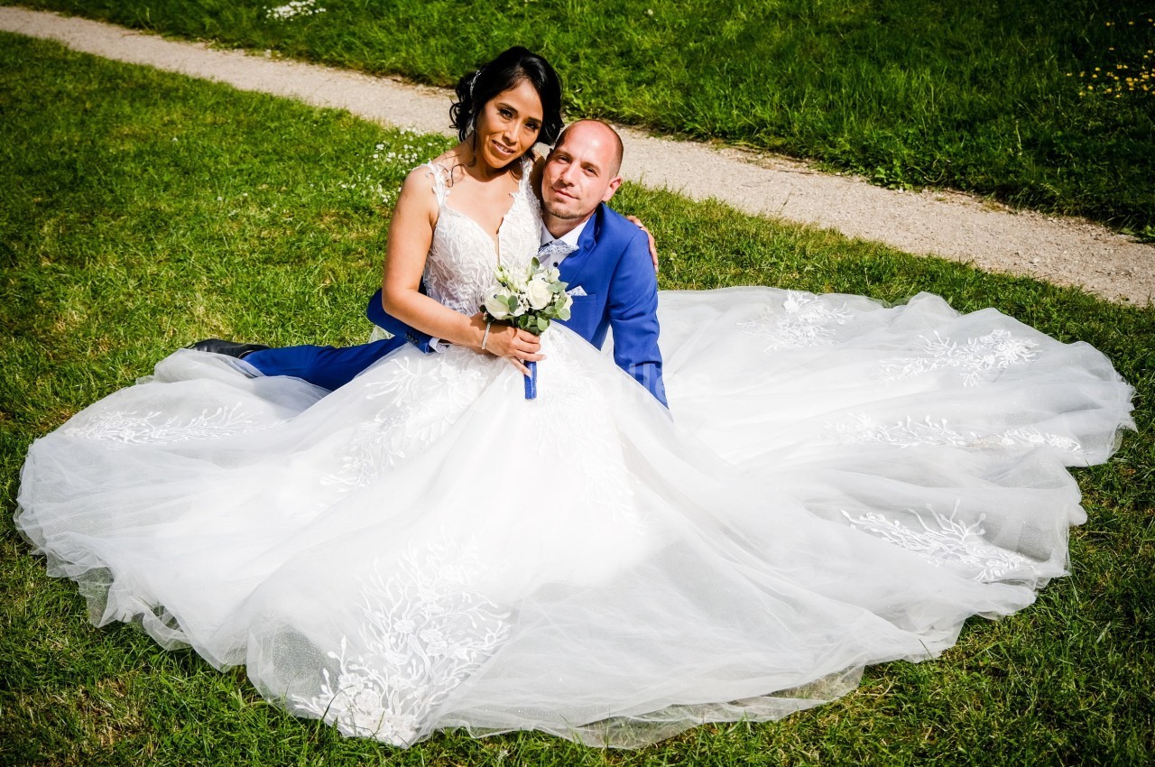 Un couple en tenue de mariage est assis sur l'herbe, la mariée portant une robe blanche volumineuse.