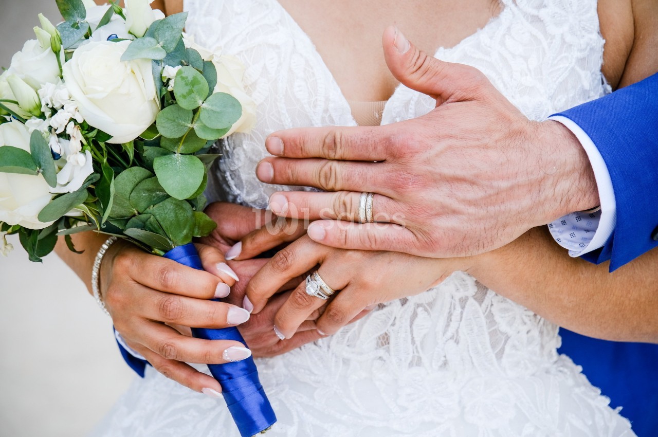 Mains d'un couple marié portant des alliances, tenant un bouquet de fleurs blanches et vertes avec un ruban bleu.