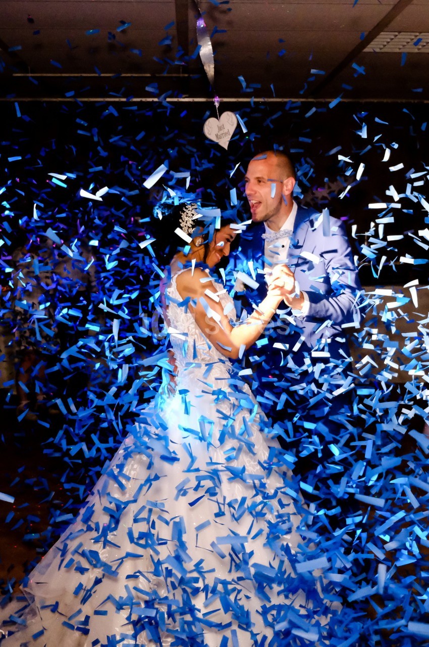 Un couple en tenue de mariage danse sous une pluie de confettis bleus dans une ambiance festive.