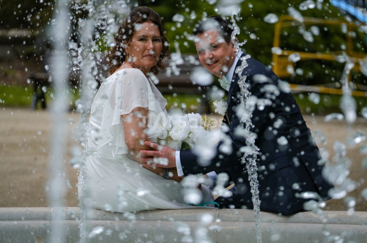 Un couple en tenue de mariage assis près d'une fontaine, avec des jets d'eau flous au premier plan.