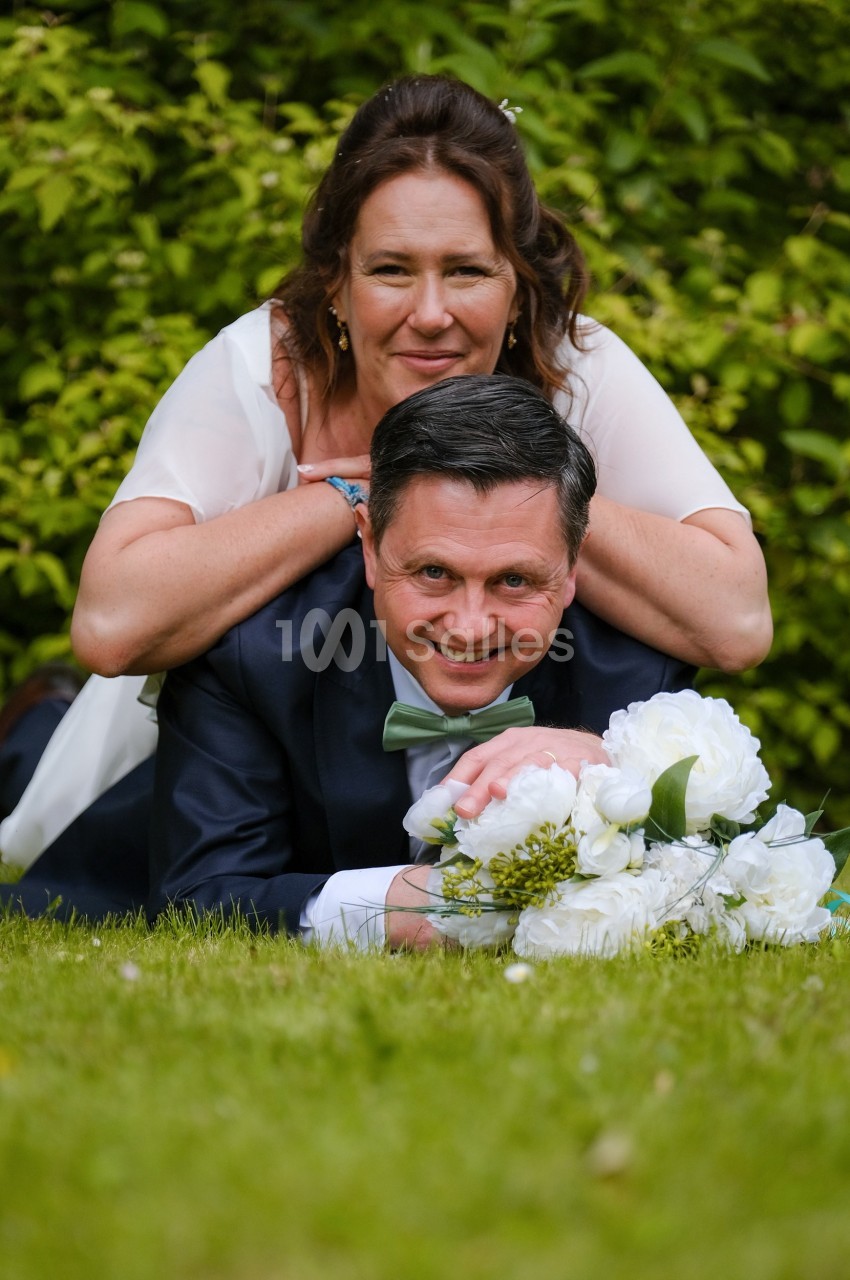 Un couple souriant allongé sur l'herbe, la femme posant ses mains sur les épaules de l'homme, avec un bouquet de fleurs…
