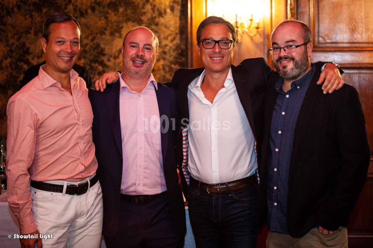 Quatre hommes souriants posent côte à côte dans un intérieur chaleureux avec un éclairage tamisé.