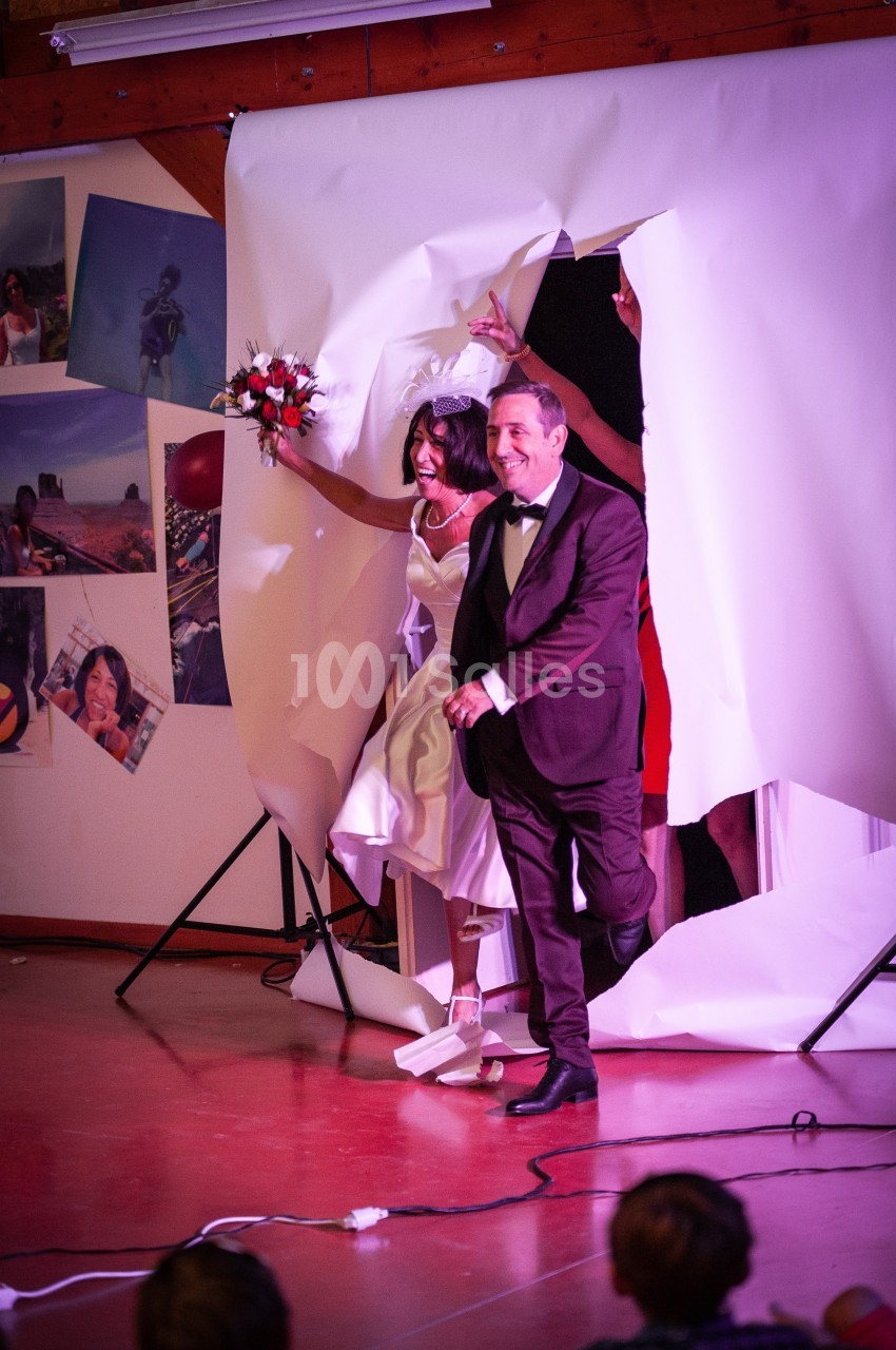 Un couple en tenue de mariage traverse une grande feuille de papier déchirée dans une salle décorée de photos.