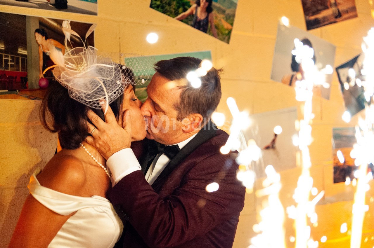 Un couple en tenue de mariage s'embrasse passionnément devant un mur décoré de photos et des étincelles.