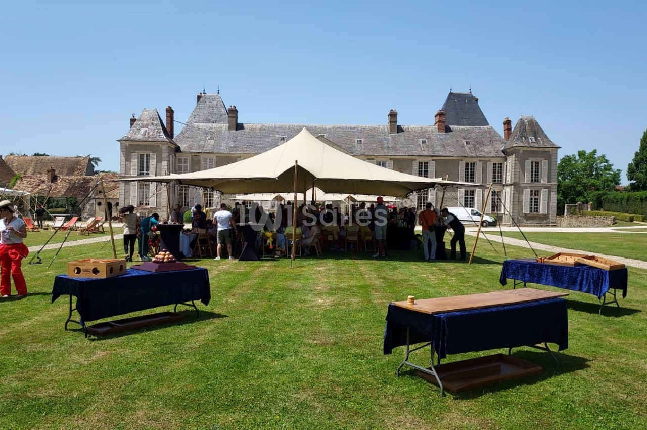 Tente dressée devant un château, entourée de tables de jeux en bois et de personnes rassemblées sur une pelouse.