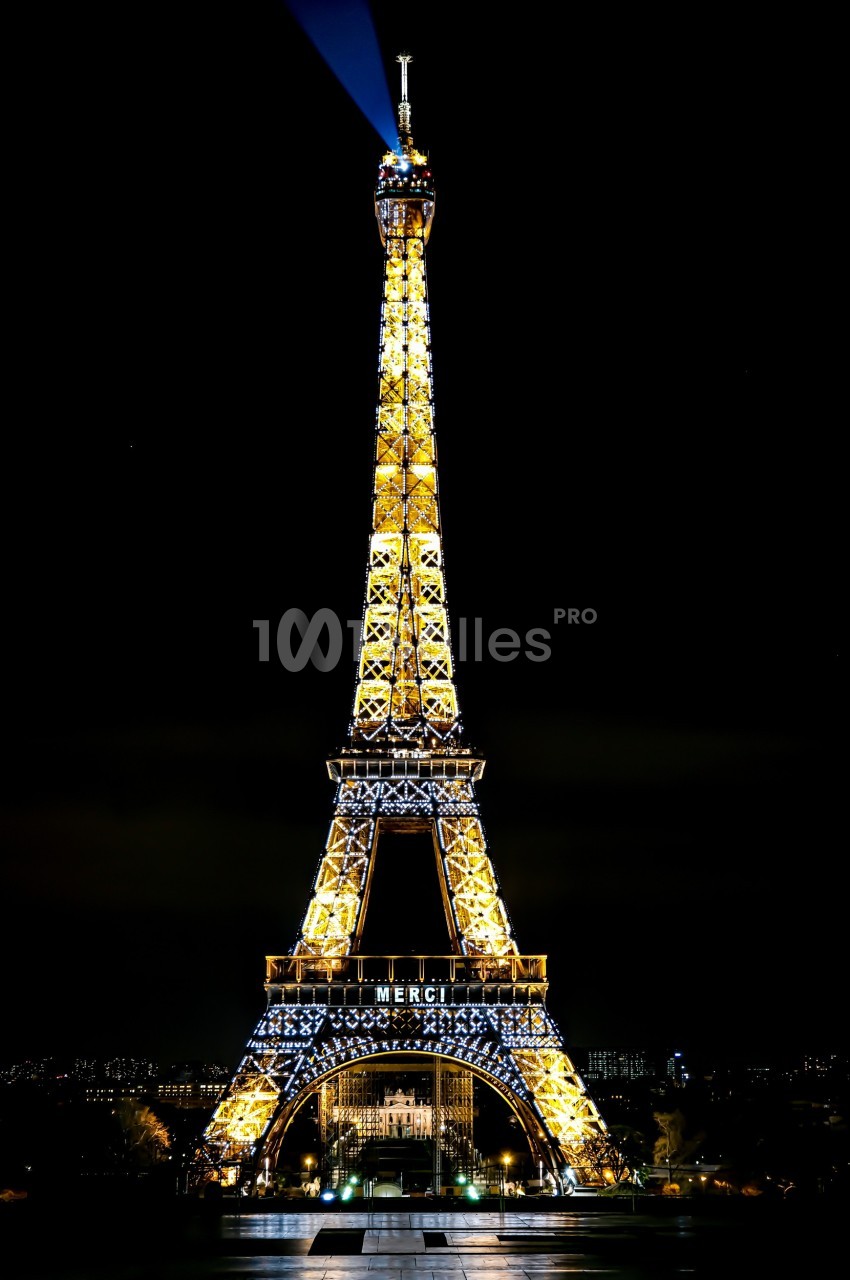 Tour Eiffel illuminée de nuit avec le mot ’MERCI’ affiché, sur fond de ciel sombre.