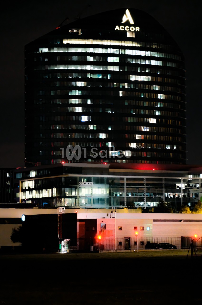 Immeuble moderne éclairé la nuit avec le logo ’Accor’ visible au sommet, entouré de bâtiments et de lumières urbaines.