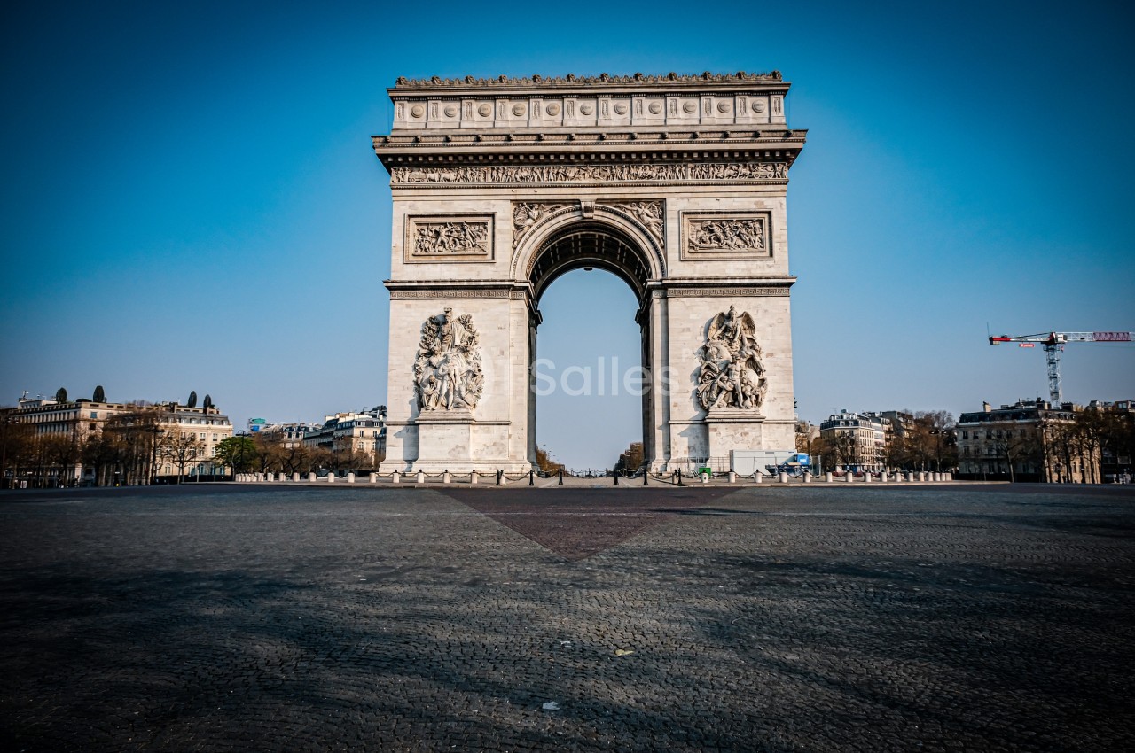 Vue frontale de l'Arc de Triomphe à Paris, entouré d'une place pavée et d'un ciel dégagé.