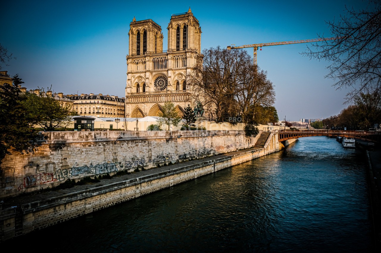 Vue de la cathédrale Notre-Dame de Paris depuis la Seine, avec un pont et une grue en arrière-plan.