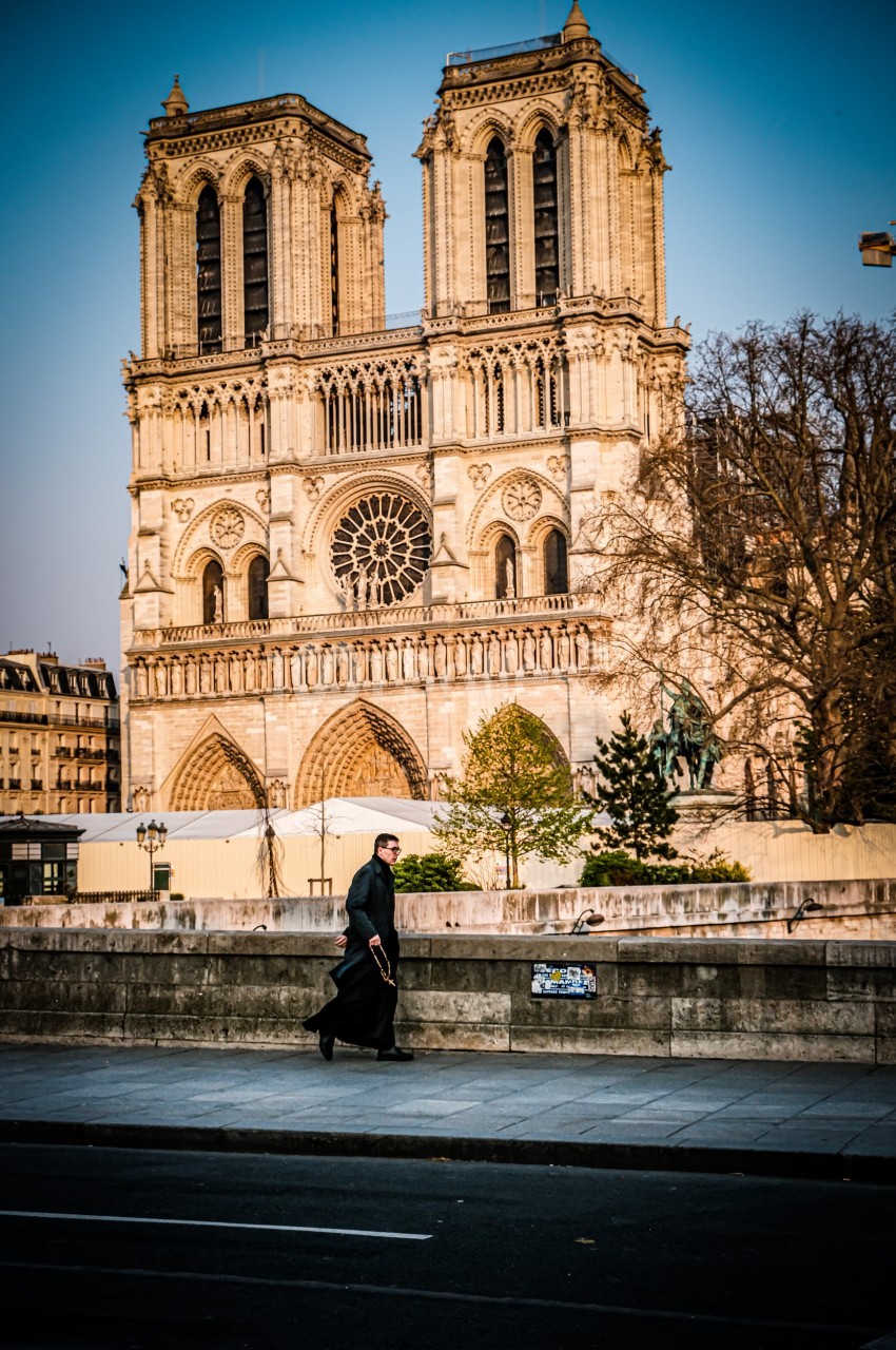 Un homme en manteau noir marche devant la cathédrale Notre-Dame de Paris par une journée ensoleillée.