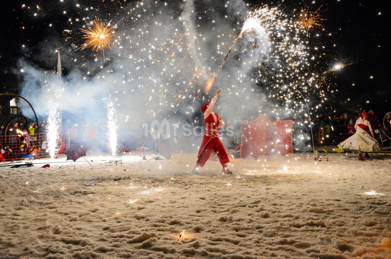 Un artiste en costume rouge manipule des bâtons enflammés dans un spectacle nocturne sur un sol enneigé.