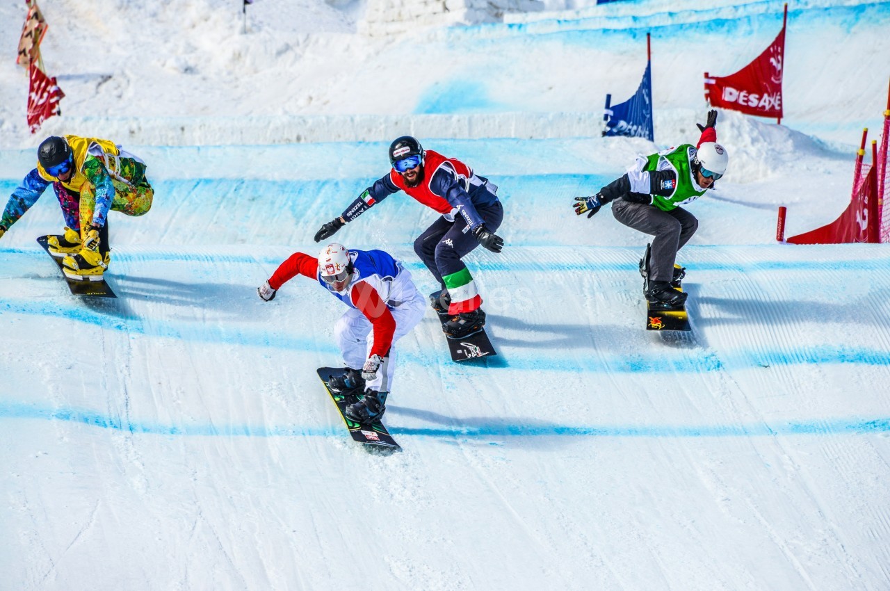 Quatre snowboardeurs en compétition descendent une piste enneigée avec des drapeaux colorés en arrière-plan.