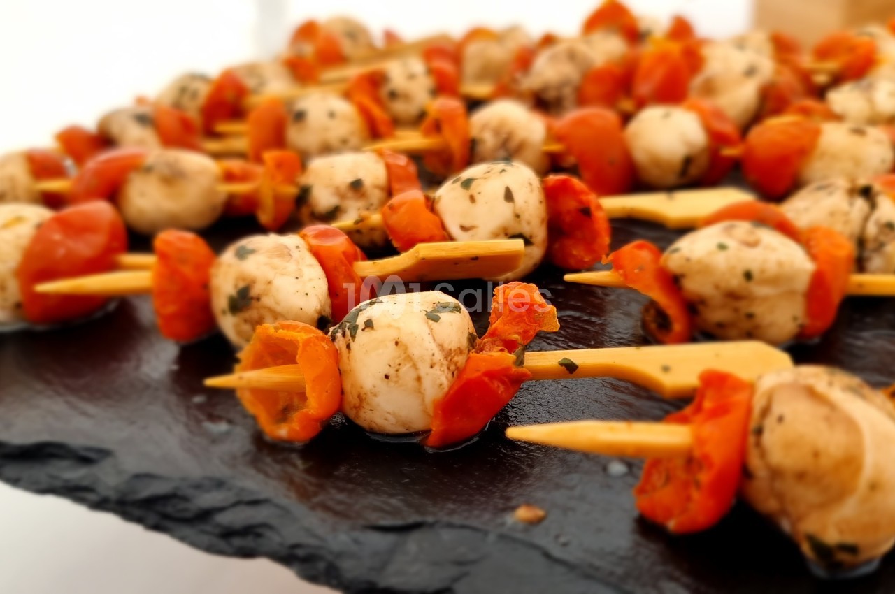Brochettes apéritives composées de tomates cerises, champignons et herbes, présentées sur une ardoise noire.