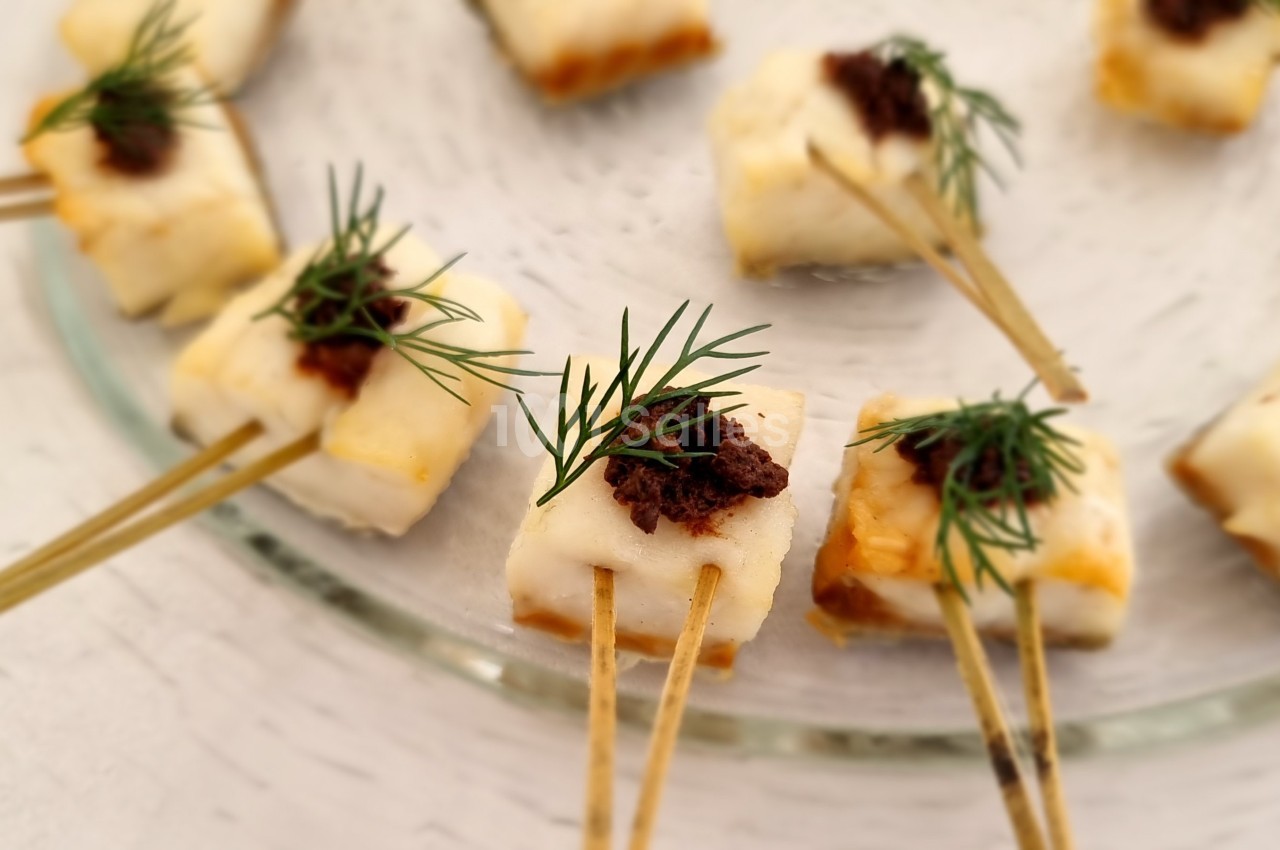 Bouchées apéritives sur des piques, composées de fromage, tapenade et brins d'aneth, disposées sur une assiette en verre.