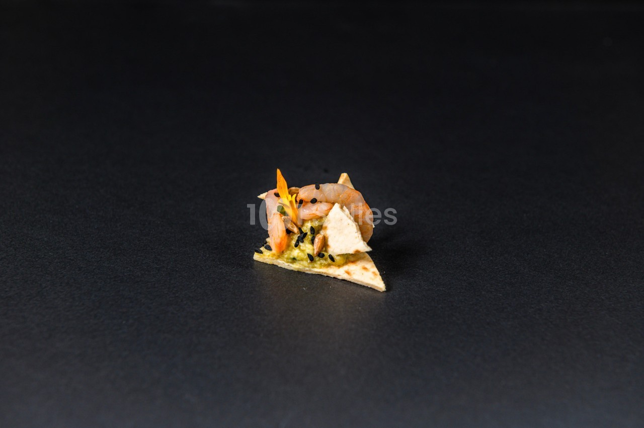 Un petit crabe posé sur un morceau de tortilla garni de guacamole, sur un fond noir.