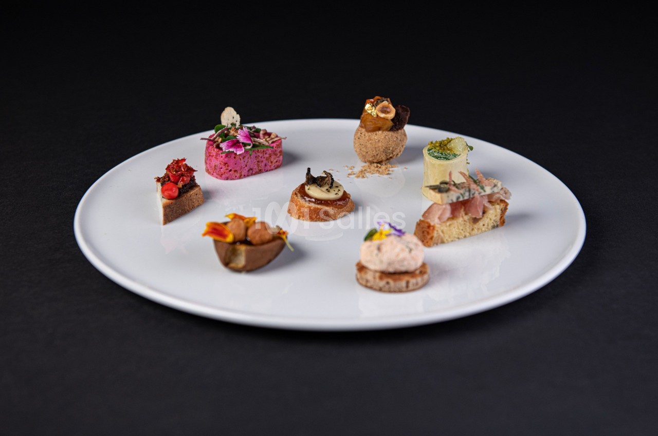 Assortiment de bouchées gastronomiques variées présentées sur une assiette blanche, sur fond noir.