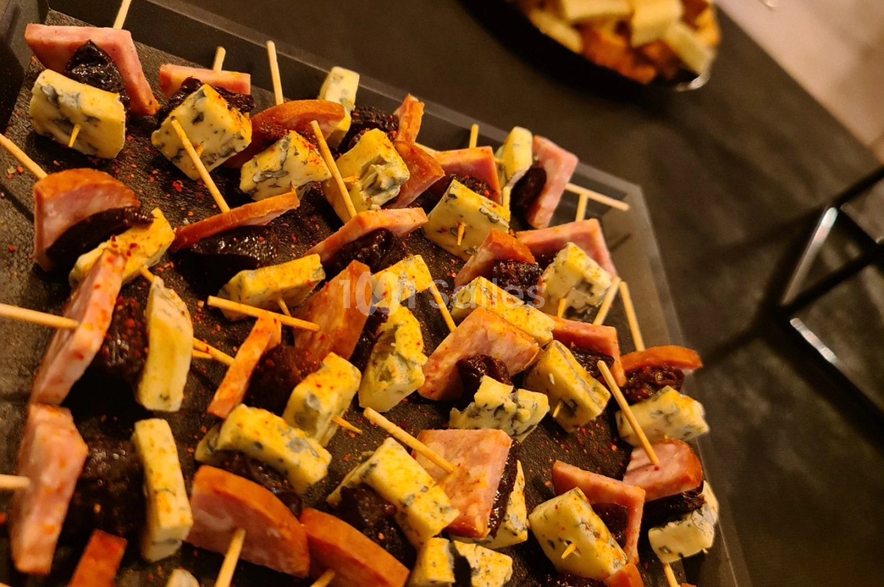 Plateau de mini-brochettes composées de fromage, saucisson et pruneaux, avec des morceaux de pain en arrière-plan.