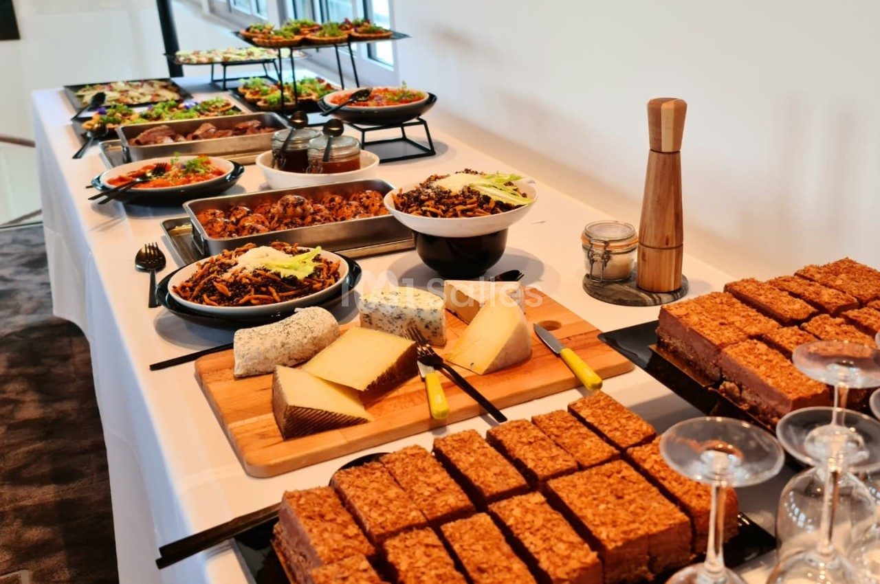 Buffet avec plats variés, fromages, desserts et couverts, disposé sur une table blanche dans un espace lumineux.