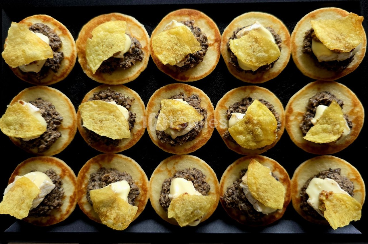 Mini-blinis garnis de tapenade, crème et chips dorées, disposés en rangées sur une surface noire.