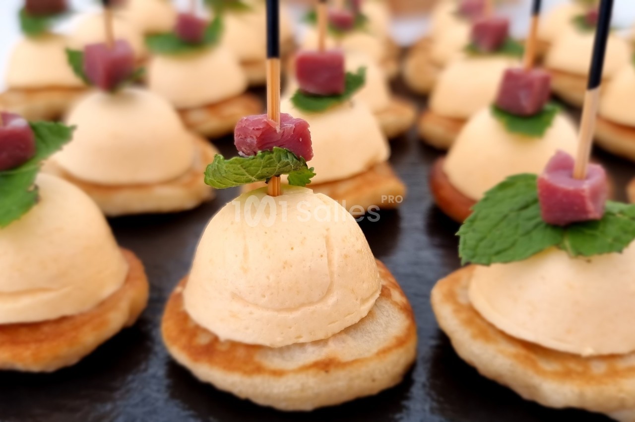 Petits canapés sur blinis garnis de mousse, feuilles de menthe et cubes de viande, présentés sur un plateau noir.