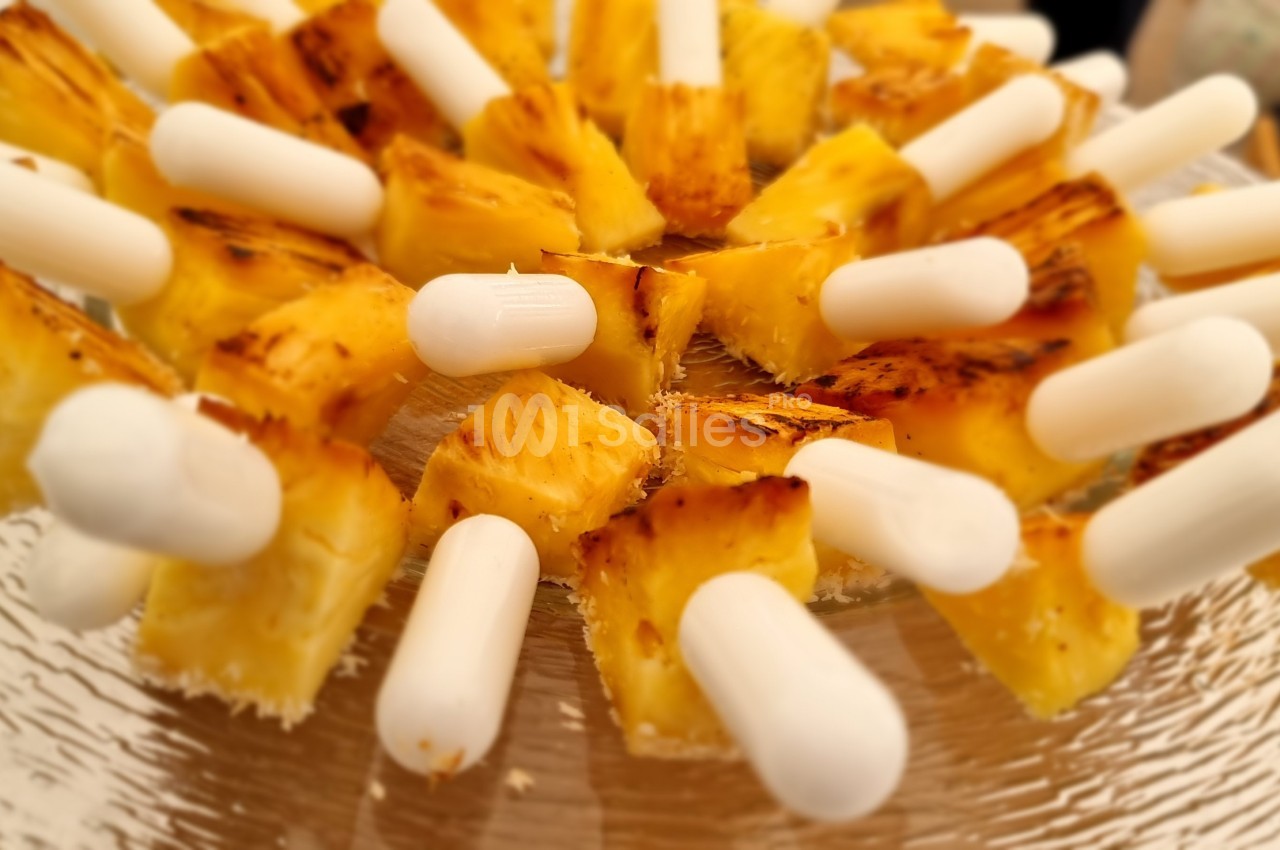 Morceaux d'ananas grillés présentés sur des bâtonnets blancs disposés en cercle sur une assiette en verre.