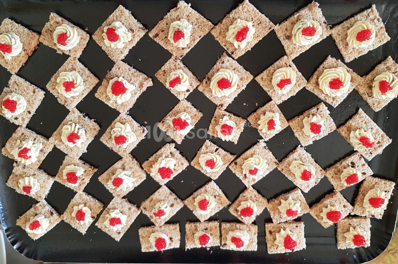 Carrés de biscuits garnis de crème et de petites décorations rouges, disposés en losanges sur un plateau noir.