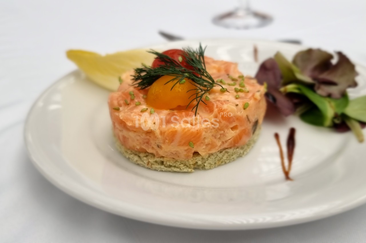 Tartare de saumon servi sur une assiette blanche avec salade verte, citron et décoration d'aneth et tomate cerise.
