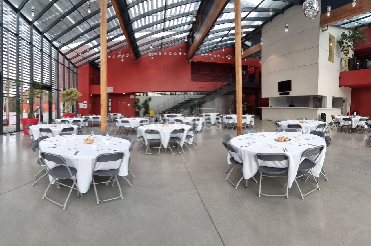 Salle spacieuse avec tables rondes dressées, nappes blanches et chaises grises, sous une verrière lumineuse.