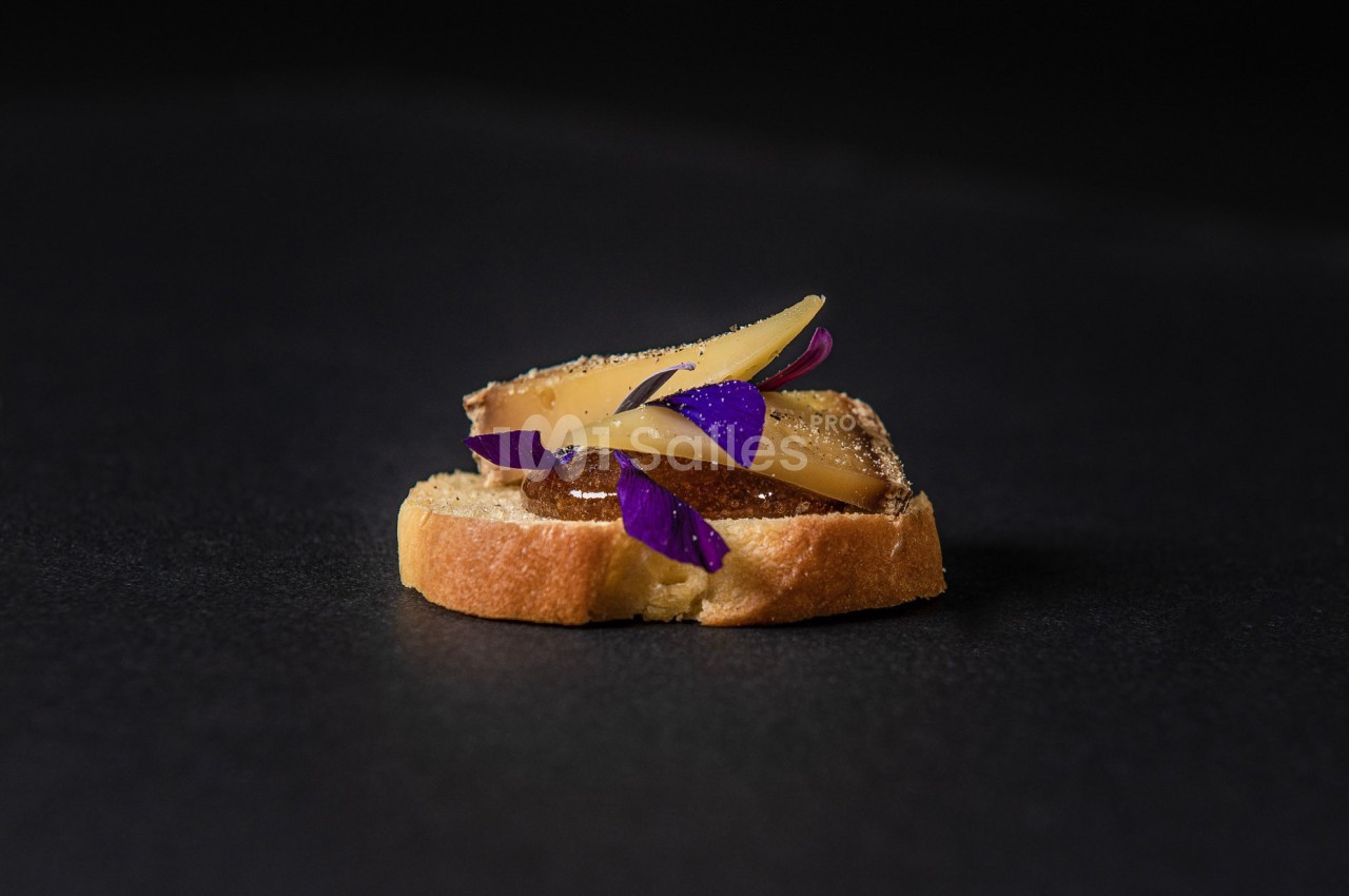 Tranche de pain garnie de fromage, fruits et pétales violets sur fond noir.