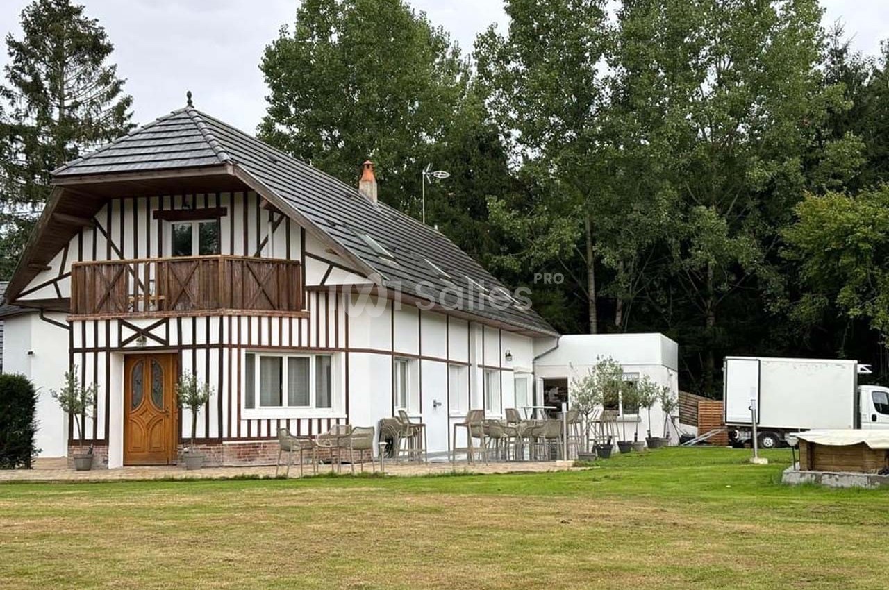 Maison à colombages avec balcon, entourée de pelouse, de tables extérieures et d'arbres en arrière-plan.