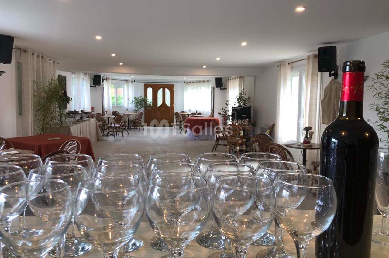 Salle de réception lumineuse avec tables dressées, chaises en osier et rangée de verres devant une bouteille de vin rouge.
