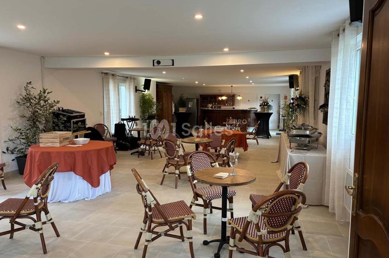 Salle lumineuse aménagée avec des tables et chaises en rotin, un buffet, et un bar en arrière-plan.
