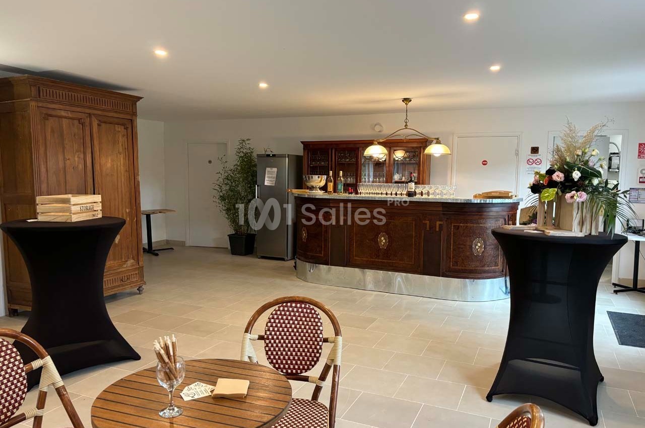 Salle lumineuse avec un bar en bois, tables et chaises en rotin, et décorations florales sur des tables hautes.