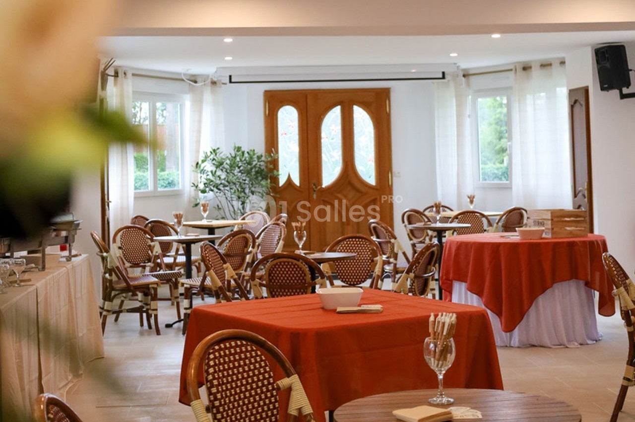 Salle lumineuse avec tables rondes et chaises en osier, nappes rouges et blanches, et grandes fenêtres donnant sur l…