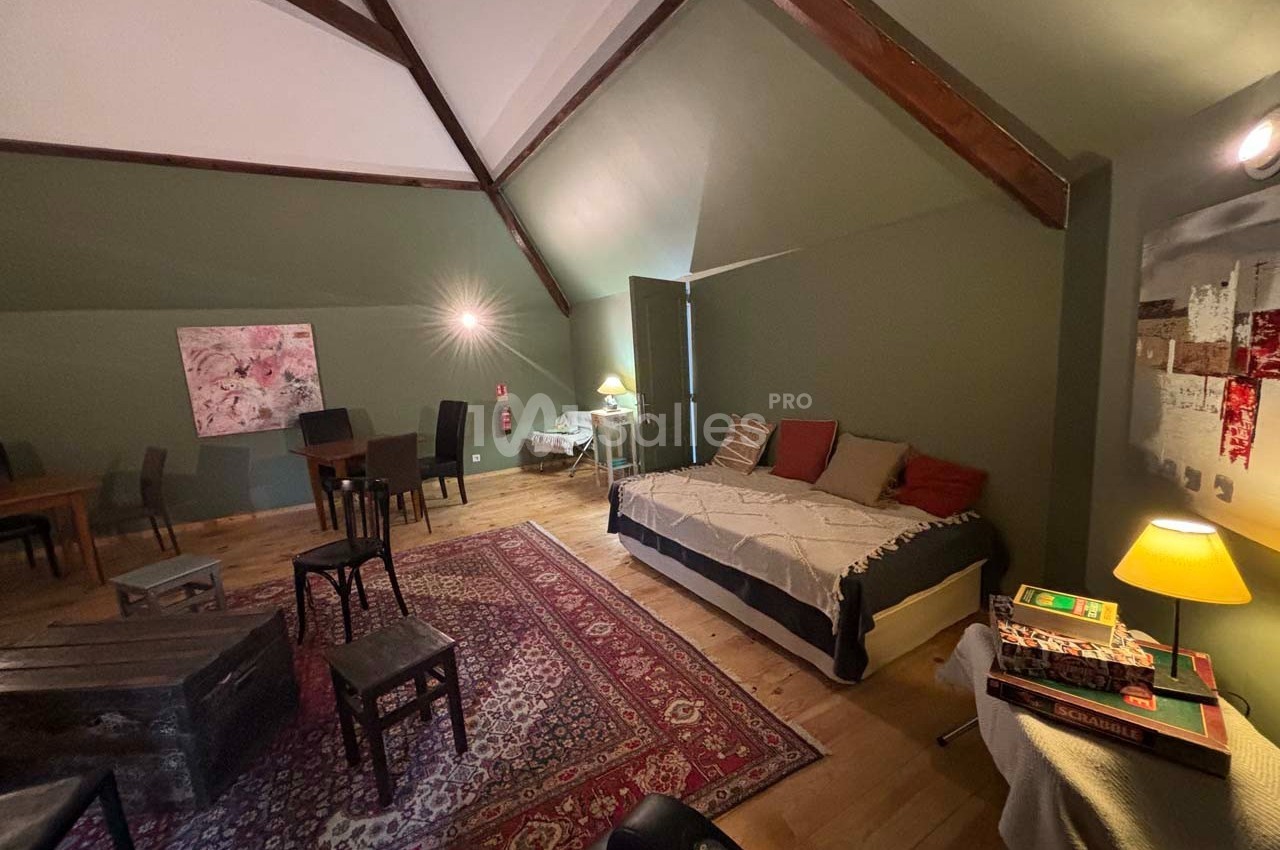 Grande pièce mansardée avec lit, tables, chaises, tapis coloré et éclairage tamisé, décorée de tableaux modernes.