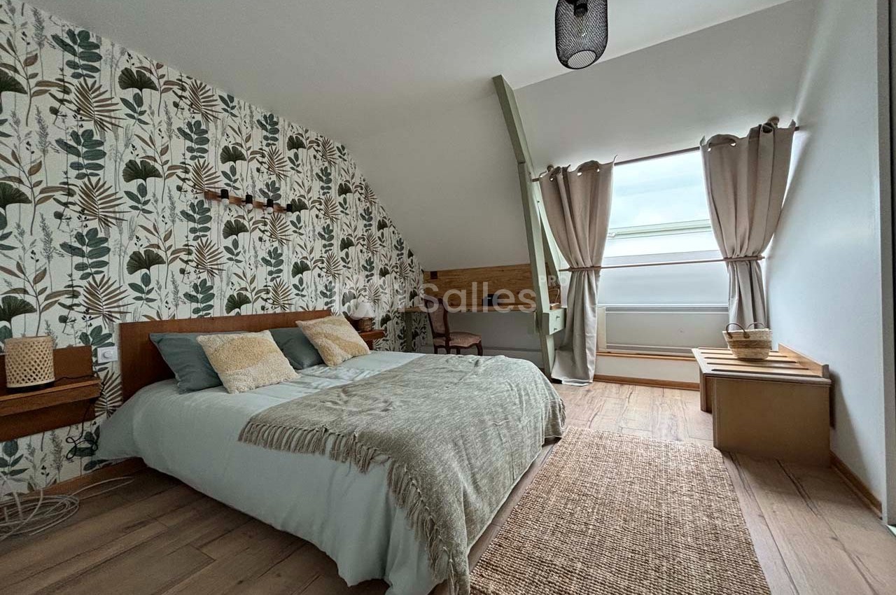 Chambre lumineuse avec lit double, décoration végétale, bureau en bois, tapis en jute et grande fenêtre avec rideaux beiges.