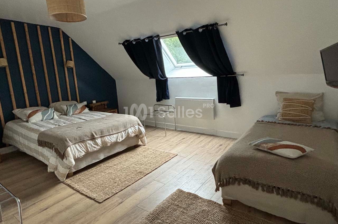 Chambre lumineuse avec un lit double, un lit simple, parquet au sol, tapis en jute et fenêtre avec rideaux noirs.