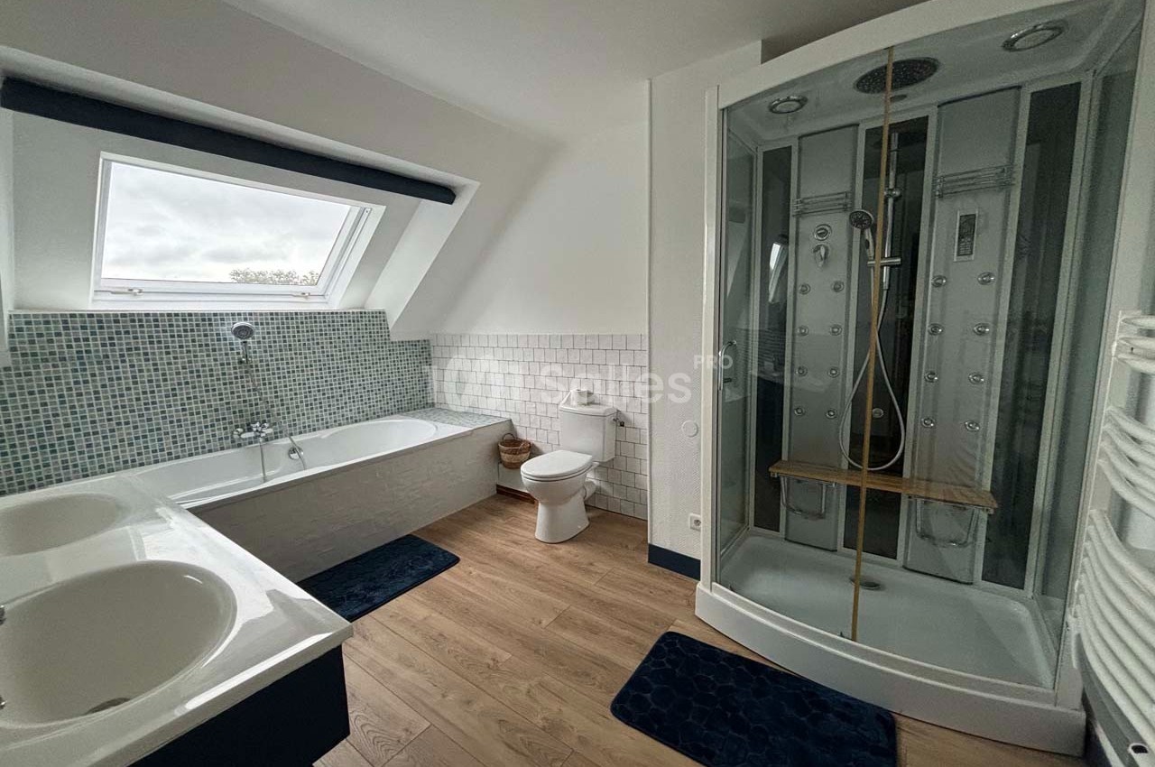 Salle de bain avec baignoire, douche à jets, lavabo double, toilettes, carrelage mural et sol en bois.