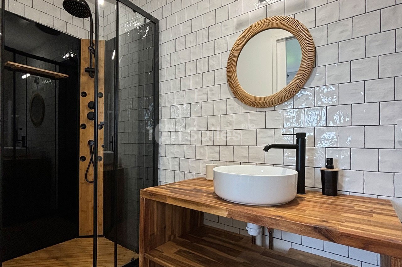 Salle de bain moderne avec douche à paroi vitrée, lavabo rond sur meuble en bois et miroir encadré en rotin.