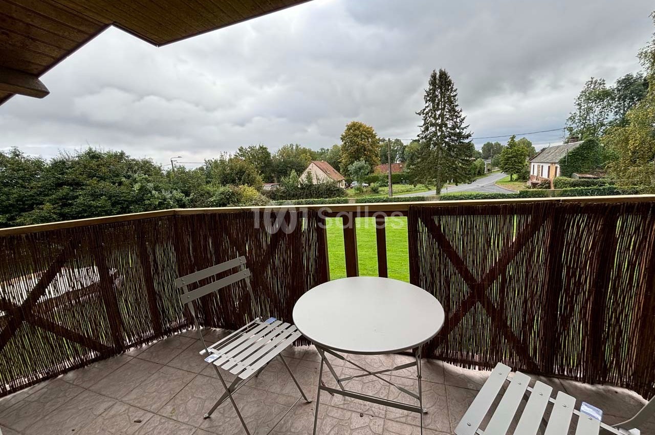 Vue depuis un balcon avec une table ronde, deux chaises blanches et un paysage rural sous un ciel nuageux.