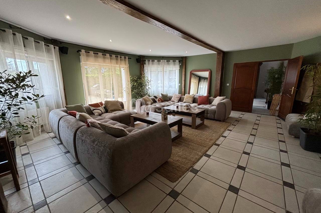 Salon spacieux avec canapés gris, table basse en bois, tapis beige et grandes fenêtres laissant entrer la lumière.
